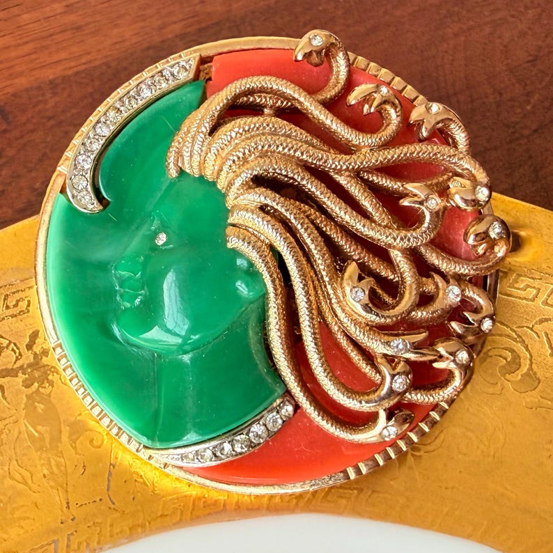 May include: A vintage brooch featuring a green face with a gold-colored snake-like hair design and red accents. The brooch is circular, with a border of clear crystals. The design is set against a gold-colored background.