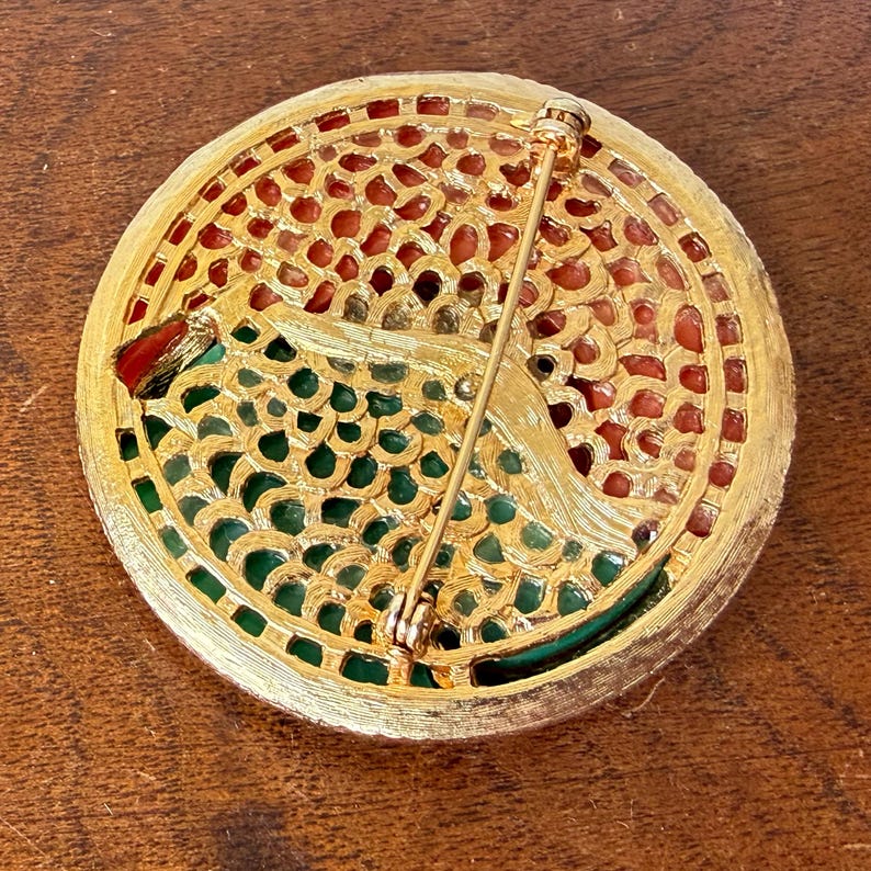 May include: A vintage gold-tone brooch with a circular design. The brooch features a filigree pattern with red and green accents. The pin is secured with a safety clasp. The brooch is likely from the mid-20th century.