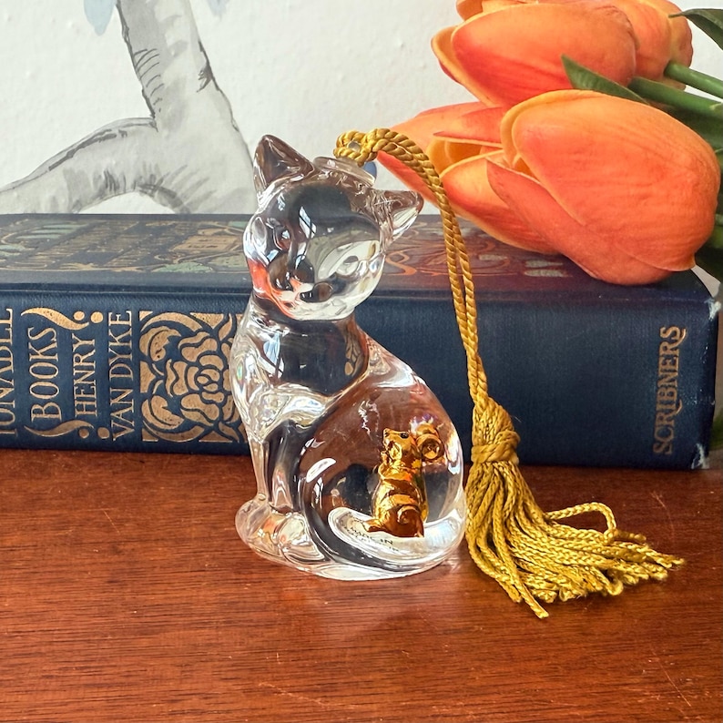 Puede incluir: Una figura de gato de cristal transparente con una borla dorada. El gato tiene una peque&ntilde;a figura de gato dorada en su interior. La figura est&aacute; sobre una superficie de madera, con un libro y tulipanes naranjas en el fondo.