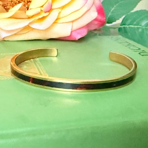 Op de afbeelding: Een goudkleurige manchetarmband met een donkerbruine en roodbruine inleg. De open armband staat tegen een groene achtergrond met een roos op de achtergrond. De sieraden hebben een eenvoudig, elegant ontwerp.