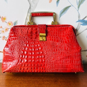 May include: A bright red handbag with a textured crocodile skin pattern. The bag features a rectangular top handle and a central vertical strap secured with a gold-toned metal clasp. The design includes a structured frame and a glossy finish.