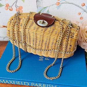 May include: A rectangular woven wicker handbag featuring a natural tan finish, a dark brown leather flap closure with a metallic turn-lock mechanism, and a gold-tone chain shoulder strap that wraps around the exterior of the bag.