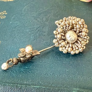 Può includere: Una spilla da bavero con un grande grappolo rotondo di perline metalliche color champagne che circondano una perla centrale liscia color bianco sporco. Lo spillo include un piccolo fiore decorativo in metallo con una minuscola perla rosa pallido.