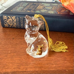Puede incluir: Una figura de gato de cristal transparente con un detalle de rat&oacute;n dorado. El gato tiene una borla amarilla atada a la cabeza. La figura est&aacute; sobre una superficie de madera, con un libro azul al fondo.