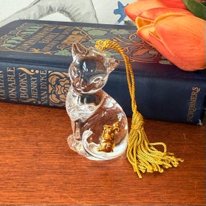 Puede incluir: Una figura de gato de cristal transparente con un rat&oacute;n dorado en su interior. El gato tiene una borla amarilla adjunta. Un libro azul con letras doradas y tulipanes naranjas en el fondo.