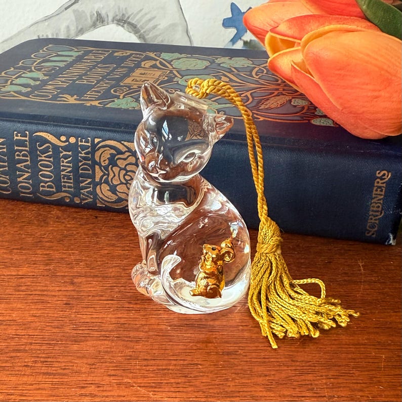 Puede incluir: Una figura de gato de cristal transparente con un rat&oacute;n dorado en su interior. El gato tiene una borla amarilla adjunta. Un libro azul con letras doradas y tulipanes naranjas en el fondo.