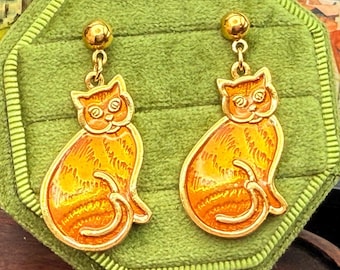 May include: A pair of gold-toned cat earrings. Each earring features a detailed orange cat design with a curled tail. The earrings are set against a green velvet background, showcasing their intricate craftsmanship.