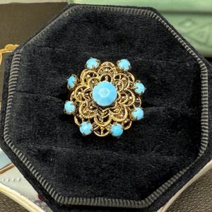 Puede incluir: Un anillo de filigrana dorado con una piedra central azul claro rodeada de piedras azules claras más pequeñas. El anillo se presenta sobre un fondo de terciopelo negro, mostrando su intrincado diseño y contraste de color.