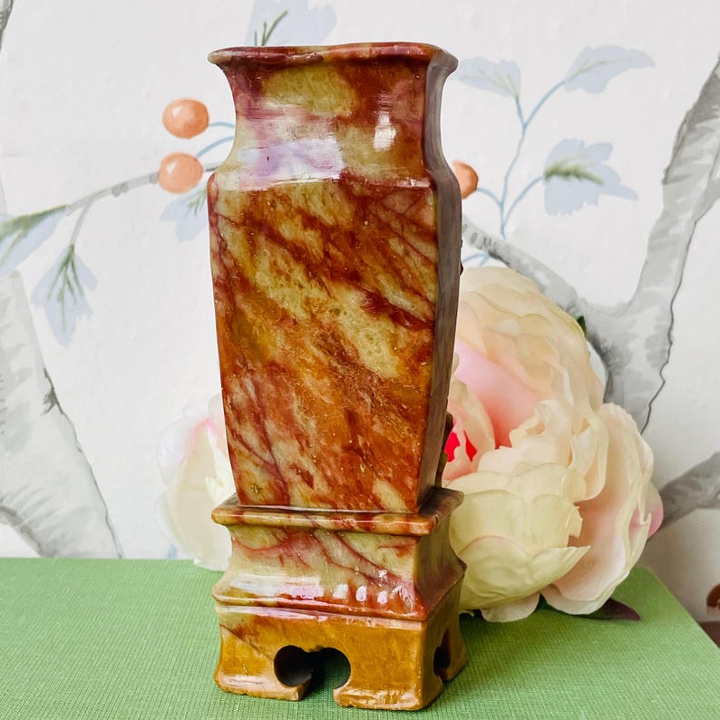 May include: A small, red and yellow marble vase with a square base and four feet. The vase is decorated with swirling patterns of red, yellow, and white.
