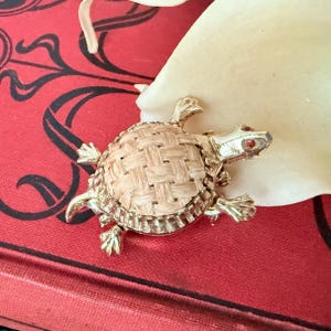 Puede incluir: Broche de tortuga dorada con caparazón de rafia tejida y ojos de piedras rojas. El broche tiene patas y cabeza detalladas, sobre un fondo rojo con detalles florales negros.