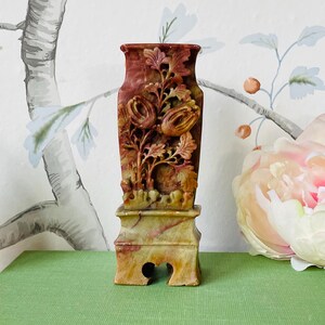 May include: A small, intricately carved stone vase with a floral design. The vase is a reddish-brown color with hints of yellow and white. It is sitting on a green surface.