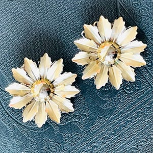 May include: A pair of vintage-style earrings. Each earring is shaped like a flower with gold-toned petals and a clear, faceted center stone. The earrings are set against a textured, dark blue background.