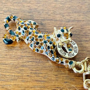 Op de afbeelding: Goudkleurige luipaardbroche met amber en zwarte kristallen accenten. De broche heeft een gedetailleerd ontwerp van een luipaard met een getextureerd oppervlak en een kettingbevestiging. De luipaard is versierd met amber en zwarte kristallen.