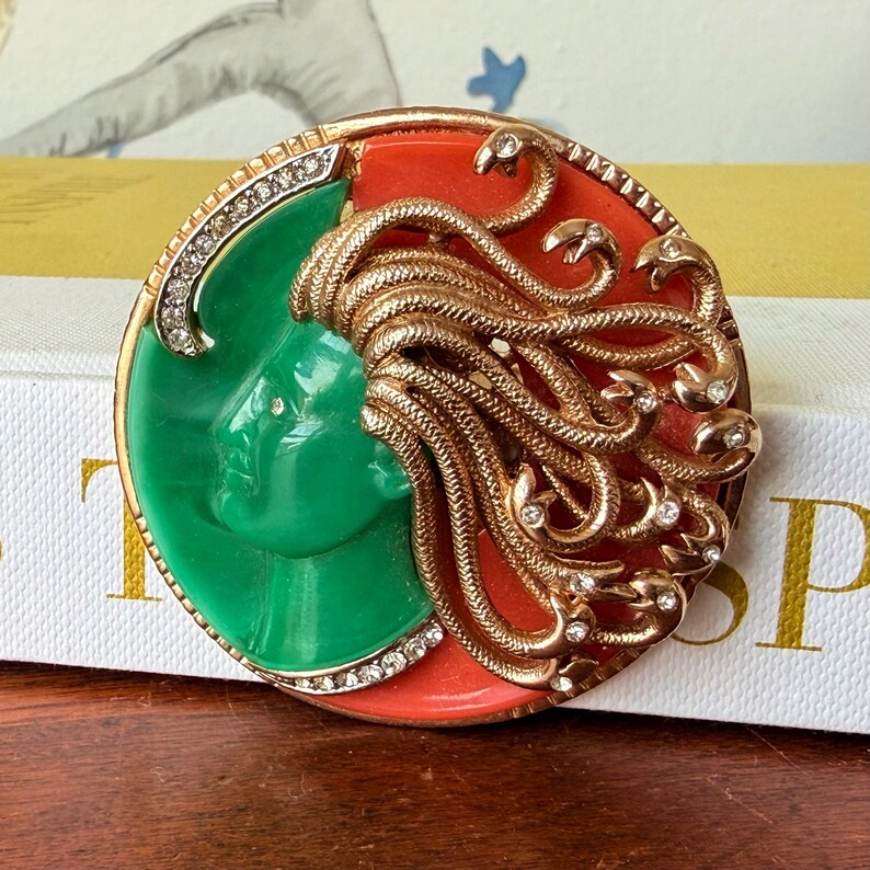 May include: A vintage brooch featuring a green face profile on a red background, encircled by a gold-tone frame with clear rhinestones. Gold-tone snakes form the hair, with additional rhinestones.