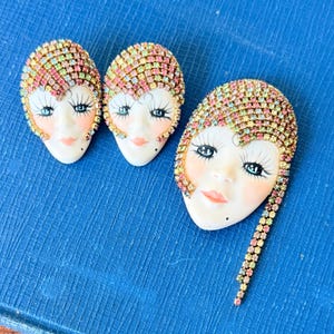May include: Three vintage face-shaped brooches with colorful rhinestone accents. The brooches feature painted faces with long eyelashes, rosy cheeks, and a beauty mark. The brooches are set on a blue background.