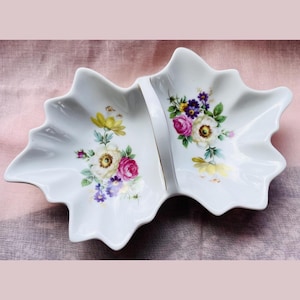 May include: White, leaf-shaped divided serving dish with floral designs. The dish features two sections separated by a white and gold stripe. Each section is decorated with colorful flowers, including yellow, pink, purple, and white blooms.