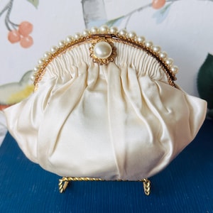 Puede incluir: Un bolso de mano de satén color crema con un diseño de tela fruncida, un marco de metal dorado y un borde decorativo de pequeñas perlas blancas a lo largo del borde superior. Una perla más grande está centrada en el cierre del bolso.