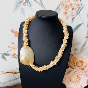 May include: A cream-colored shell necklace displayed on a black mannequin. The necklace features a large, oval pendant and a strand made of small, irregular shell pieces. The background includes a floral wallpaper and a few pink roses.