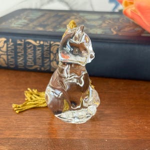 Puede incluir: Una figura de gato de cristal transparente con una borla dorada. El gato est&aacute; sentado, con la cabeza girada hacia un lado. La figura est&aacute; sobre una superficie de madera, con un libro en el fondo.