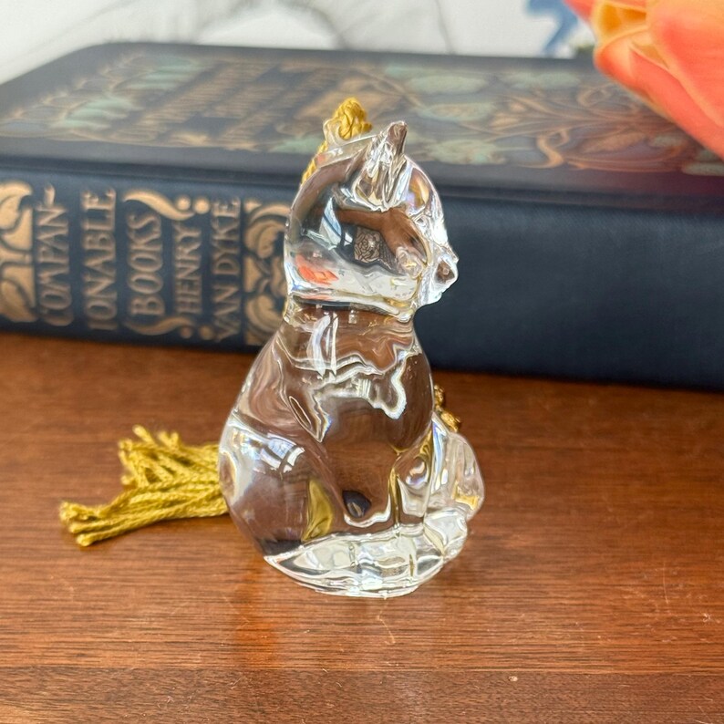 Puede incluir: Una figura de gato de cristal transparente con una borla dorada. El gato est&aacute; sentado, con la cabeza girada hacia un lado. La figura est&aacute; sobre una superficie de madera, con un libro en el fondo.