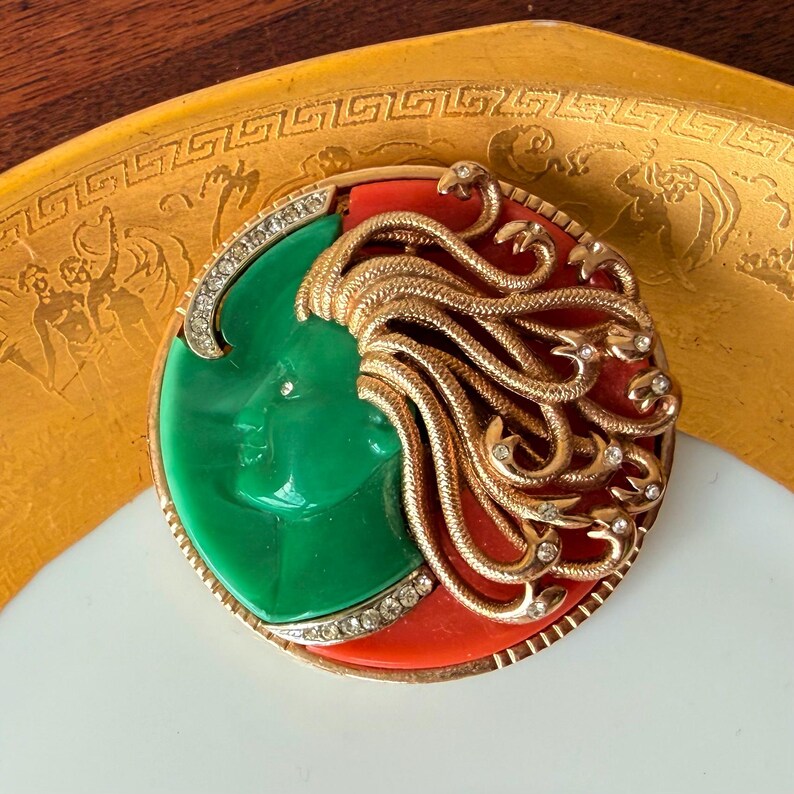 May include: A vintage brooch featuring a green face with flowing gold hair, set against a red background. The brooch is framed with a gold border and encrusted with small, sparkling crystals. The design is circular and ornate.