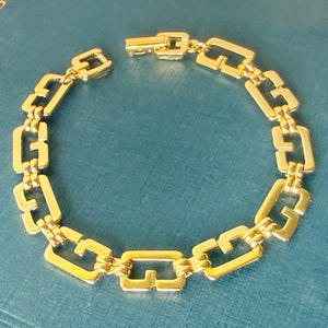 May include: A gold-coloured bracelet featuring a repeating rectangular link design. The bracelet is made of metal and has a clasp closure. The links are connected by smaller, decorative links. The bracelet is shown on a blue surface.