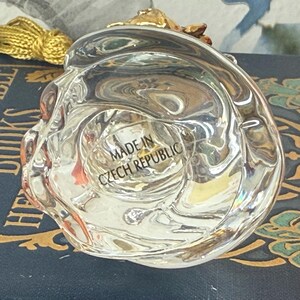 Puede incluir: Un objeto de vidrio transparente y esculpido con un acento dorado. La base del objeto es visible, con el texto "MADE IN CZECH REPUBLIC" grabado en el vidrio. El objeto es probablemente una pieza decorativa o un peque&ntilde;o recipiente.