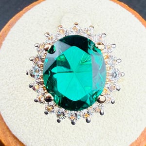 May include: A ring featuring a large, oval-cut, vibrant green centre stone surrounded by a halo of numerous small, clear, round-cut stones. The setting is metallic, and the stones are arranged in a circular, floral-like pattern on a light base.