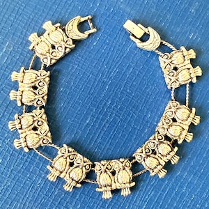 May include: Silver-toned owl bracelet with a decorative clasp. The bracelet features multiple owl figures linked together with a delicate chain. The owls are detailed with feathers and eyes, creating a vintage aesthetic. The bracelet is shown on a blue surface.