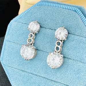 May include: A pair of silver-toned drop earrings featuring two round, clear, faceted stones connected by a figure-eight shaped link. The upper stone is smaller than the lower stone, and both are set in a polished metal frame with a secure backing.