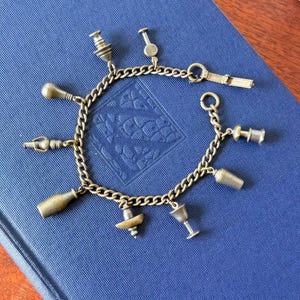 May include: A vintage charm bracelet with a gold-toned chain and various charms. The charms include miniature representations of bar tools, such as a cocktail shaker, bottle, and corkscrew. The bracelet is displayed on a blue surface.