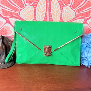 以下が含まれることがあります： ゴールドカラーの金具が付いた、明るい緑色の封筒型クラッチバッグ。クラッチバッグは、テクスチャ加工された表面、折り返しフラップ、リストストラップが特徴です。留め金は、ブランド名「Lulu」が刻印されたゴールドカラーの金属です。