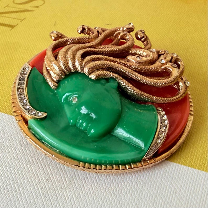May include: A vintage brooch featuring a green Medusa head with gold snakes and a red background. The brooch has a gold frame with rhinestone accents. A unique and eye-catching piece of jewelry.