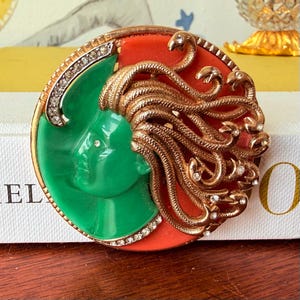 May include: A vintage brooch featuring a Medusa design. The brooch has a green face, red background, and gold-colored snakes for hair. The brooch is round and has a gold-colored border with clear rhinestones.