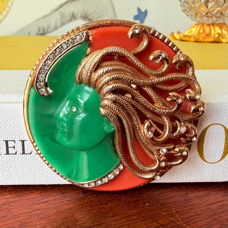 May include: A vintage brooch featuring a Medusa design. The brooch has a green face, red background, and gold-colored snakes for hair. The brooch is round and has a gold-colored border with clear rhinestones.