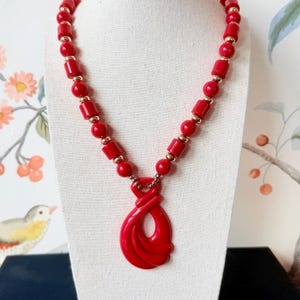 May include: A red beaded necklace with a pendant. The necklace features a series of red beads in various shapes, interspersed with small gold-colored beads. The pendant is a large, abstract red shape. The necklace is displayed on a white mannequin.