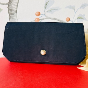 May include: A rectangular navy blue clutch bag with a smooth fabric finish. The front flap features a single centered circular gold-toned button accent with a white pearl-like inlay. The bag has a structured shape with angled bottom corners.