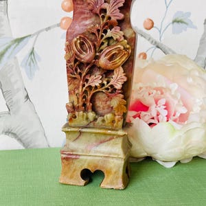 May include: A carved stone vase with a floral design. The vase is a light green and brown color with red accents. It has a square base with a carved design.