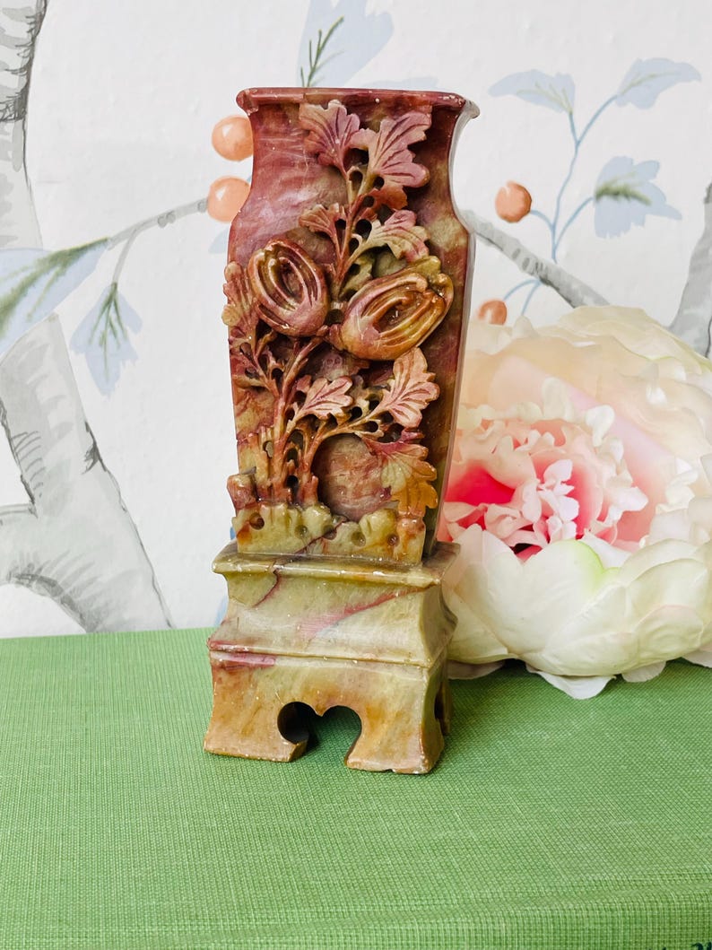 May include: A carved stone vase with a floral design. The vase is a light green and brown color with red accents. It has a square base with a carved design.