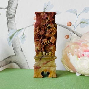 May include: A carved stone vase with a floral design in shades of red, brown, and green. The vase is on a pedestal with a carved base.