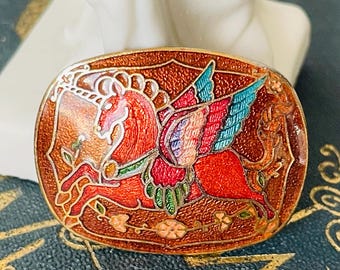 May include: A red, winged unicorn with a gold horn and blue wings is depicted on a brown, oval-shaped brooch. The brooch is decorated with gold and green accents and has a textured surface.