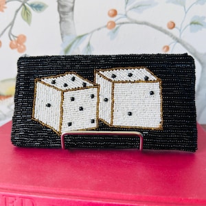 May include: A rectangular clutch bag covered in black seed beads featuring a beaded graphic of two white dice with gold-toned outlines and black pips. The bag has a structured rectangular shape and is displayed on a small metal stand.