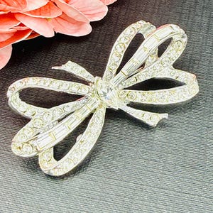 May include: A silver-toned metal brooch shaped like a bow, featuring a central marquise-cut clear crystal and multiple baguette-cut crystals along the center. The loops of the bow are encrusted with numerous small, round, clear rhinestones.