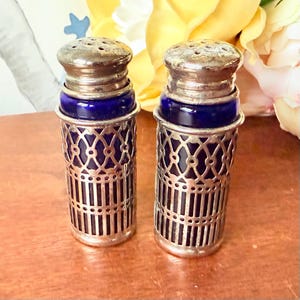 May include: A pair of antique salt and pepper shakers. Each shaker has a cobalt blue glass bottle encased in a silver filigree holder. The tops are silver with small holes for dispensing.