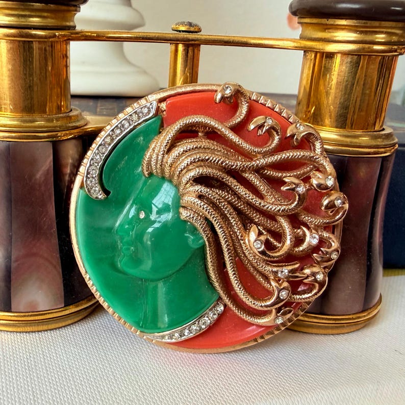May include: A round, ornate brooch featuring a green face on a red background, with gold-colored snakes and clear crystals. The brooch is set in a gold-colored frame with a diamond-like border. The background includes a pair of vintage binoculars.