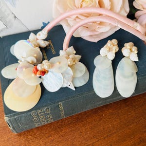 May include: A necklace and matching earrings set featuring iridescent shell-like elements, pearls, and coral-colored beads. The necklace has a pink satin ribbon. The jewelry is displayed on a vintage book.
