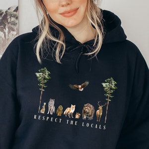 K&ouml;nnte beinhalten: Schwarzer Kapuzenpullover mit einem Aufdruck verschiedener Tiere in einer Waldlandschaft. Der Text "Respect the Locals" ist unter den Tieren gedruckt.