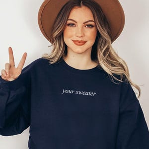 Può includere: Una felpa blu navy con la scritta ricamata "your sweater" in un carattere bianco e delicato. La modella indossa un cappello marrone e fa il segno della pace con le dita.