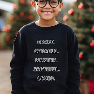 May include: A young person wearing a black sweatshirt with the words "BRAVE. CAPABLE. WORTHY. GRATEFUL. LOVED." printed in white.