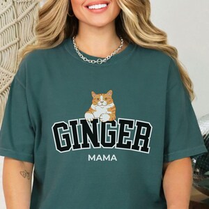 May include: Teal blue t-shirt with a graphic of an orange tabby cat and the text "GINGER MAMA" in black block letters.